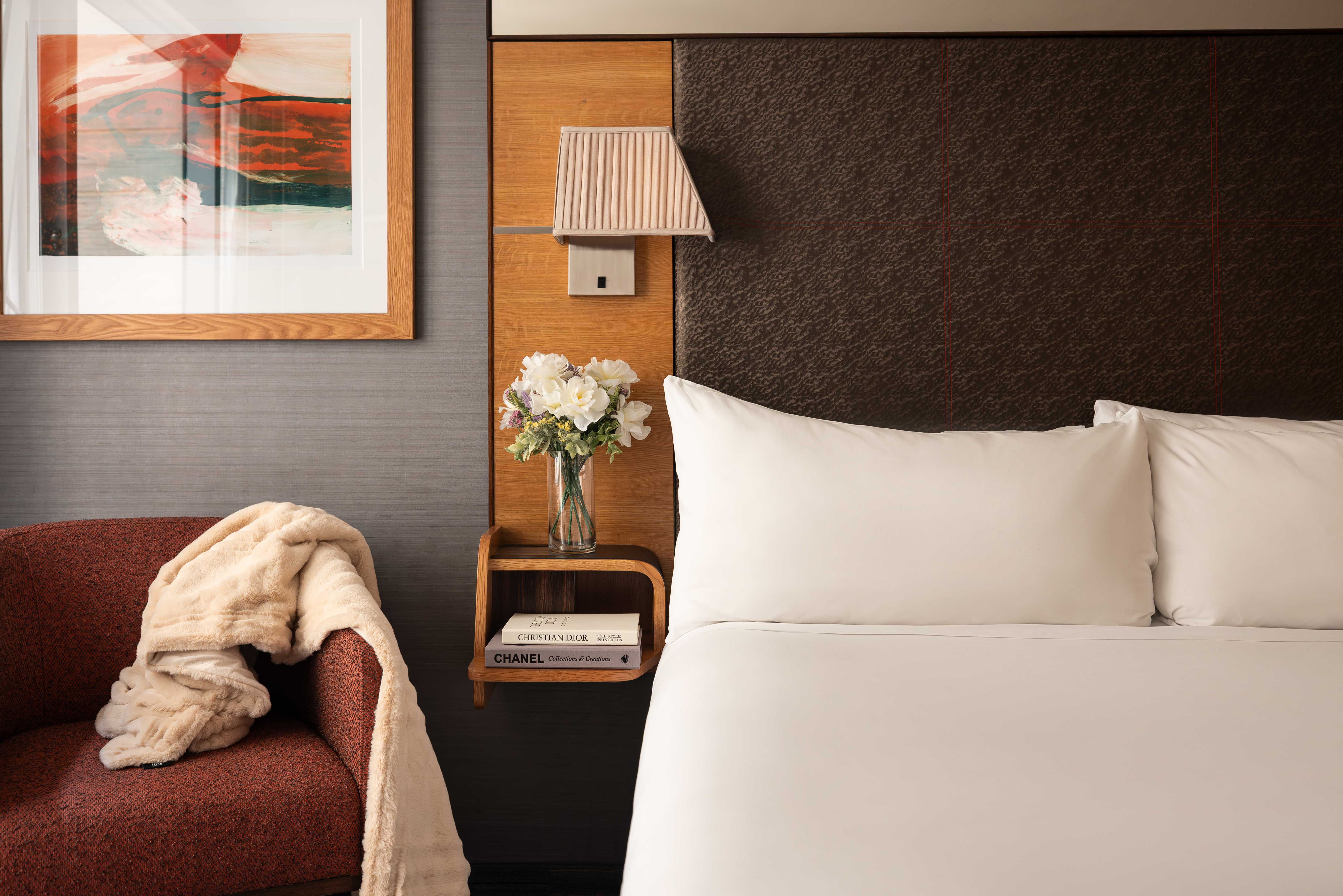 A hotel room with a bed, chair, lamp, and flowers.